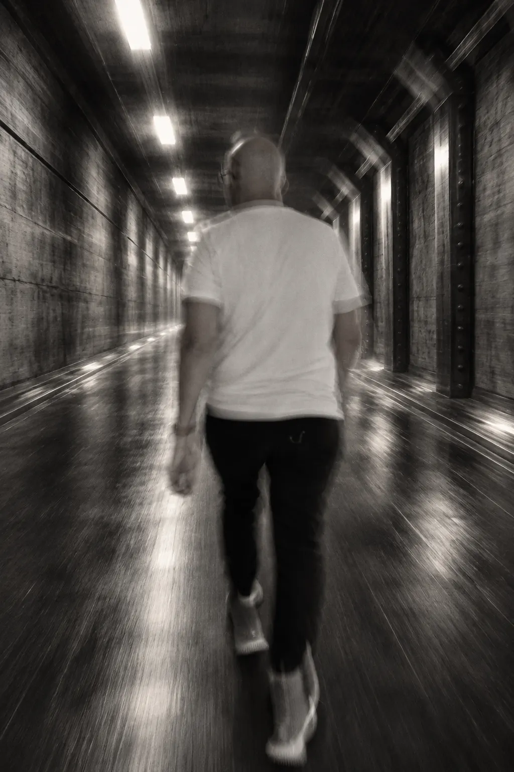 Nick Dalk de dos marche dans un couloir industriel, sa silhouette se fondant dans le mouvement.